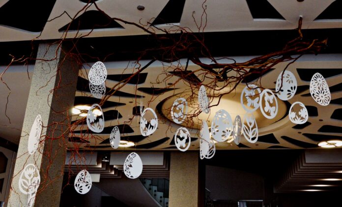 DSC_2740 Pod ozdobnym sufitem w holu jasielskiego domu kultury wiszą przyczepione do gałęzi ręcznie robione pisanki z białego papieru. Obok szary filar, w tle widoczne światła sufitowe