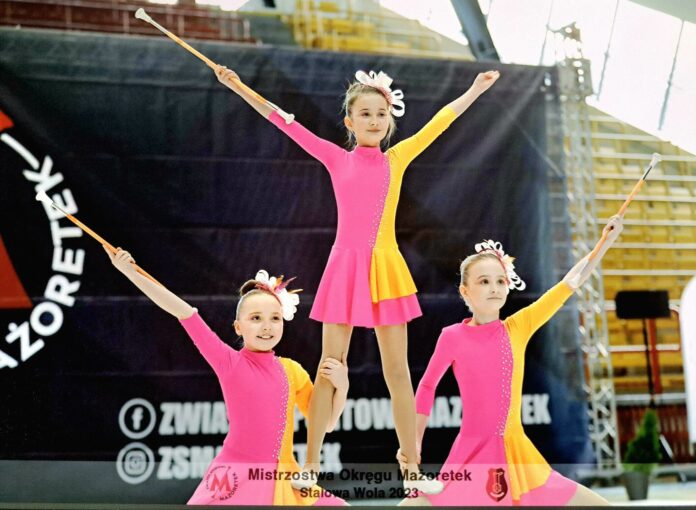 Mażoretki 1 Obraz zawiera: Trzy dziewczynki w strojach mażoretek w wieku około 9 lat. Dwie z nich jedną ręką podtrzymują stojącą na ich udach koleżankę, która unosi obie ręce w gorę. W prawej ręce trzyma batutę. Podobnie dwie jej koleżanki. Jedna trzyma swoją batutę w prawej, druga w lewej ręce. W ten sposób tworzą typową figurę piramidy.