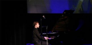 Odkrywanie muzyki Chopina w JDK Obraz zawiera: Młody mężczyzna siedzi bokiem do widowni na scenie JDK. Przed nim fortepian i mikrofon statywie. Trzyma ręce na klawiszach fortepianu. Z tyłu nad sceną wisi duży ekran na którym widać ręce pianisty.