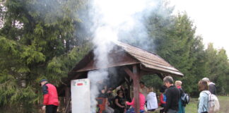 Zwiedzanie z KK-T JDK i PTTK: Odludne ścieżki bieszczadzko-beskidzkie Na tle polany stoi drewniana wiata, pod nią, na ławkach siedzą ludzie przy posiłku. Obok wiaty pali się ognisko. W tle duże choinki