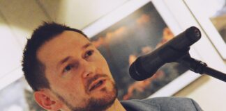 Magicznie i majestatycznie w Galerii JDK Marcin Bawiec, autor wystawy stoi przed mikrofonem i opowiada o swoich górskich wyprawach. Na sobie ma niebieską marynarkę, grantową koszulkę z czarnym nadrukiem. Ma krótkie czarne włosy i krótki czarny zarost na brodzie. W rękach trzyma teczkę