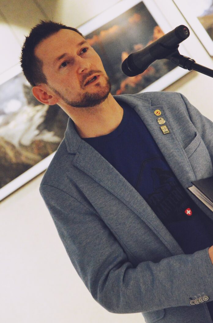 Marcin Bawiec, autor wystawy stoi przed mikrofonem i opowiada o swoich górskich wyprawach. Na sobie ma niebieską marynarkę, grantową koszulkę z czarnym nadrukiem. Ma krótkie czarne włosy i krótki czarny zarost na brodzie. W rękach trzyma teczkę