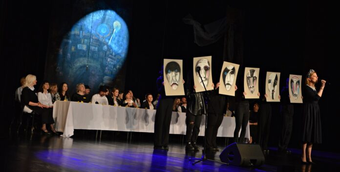 DSC_3025 Na scenie znajduje się kilka postaci ubranych na czarno, trzymają przed sobą w dłoniach namalowane na teksturze maski. Przed nimi stoi statyw na mikrofon, przed nim leży duży głośnik, a nieco dalej stoi młoda kobieta, blondynka z wiankiem na głowie i śpiewa do mikrofonu. Ma krótkie włosy, czarną sukienkę z białym kołnierzykiem oraz czarne buty na wysokim obcasie. Z tyłu stoi długi stół przykryty białym obrusem, siedzą przy nim dorośli ludzie ubrani na białoczarno.. Za nimi, na ciemnym tle podświetlona okrągła niebieska dekoracja z motywem domów