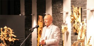 Obraz zawiera: Na pierwszym planie przed mikrofonem na statywie stoi łysy mężczyzna w wieku około 60 - ciu lat. Dłonie złączył na wysokości przepony i przemawia do przybyłych na spotkanie. Z tyłu za nim stoją jego prace, podświetlone od góry punktowymi lampami ściennymi.