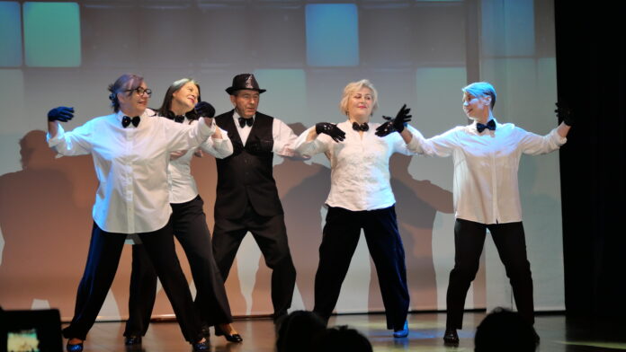 Na tle jasnego ekranu tańczą seniorzy jasielskiego domu kultury wraz z instruktorką tańca jasielskiego domu kultury. Cztery panie mają białe koszule i czarne muszki oraz czarne spodnie. Za nimi mężczyzna w czarnym kapeluszu na głowie, białej koszuli, z czarną muszką pod szyją, na koszuli ma czarną kamizelkę oraz czarne spodnie. Wszyscy mają na dłoniach czarne rękawiczki. Trzy panie mają jasne włosy, jedna dłuższe ciemne