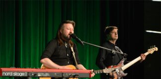 Obraz zawiera: Na scenie Jasielskiego Domu Kultury stoją dwaj mężczyźni z gitarami w dłoniach na których grają. Pierwszy od lewej jest szatynem z dłuższymi włosami do ramion. Gra na gitarze elektrycznej, śpiewa do mikrofonu, przed nim stoją również klawisze. mężczyzna z prawej stronie gra na gitarze basowej.