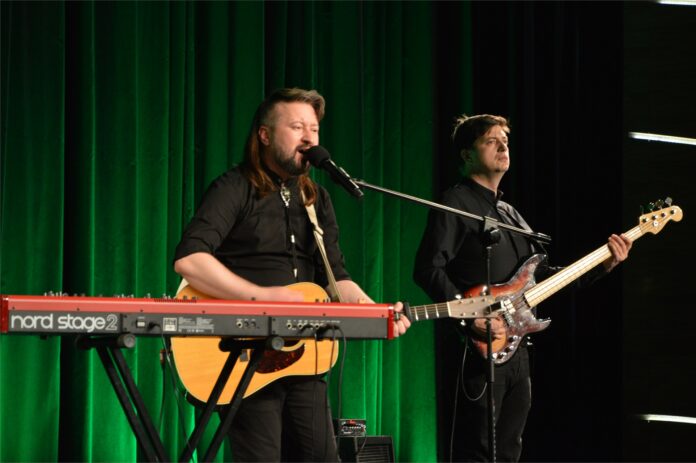 Piramidy_4 Obraz zawiera: Na scenie Jasielskiego Domu Kultury stoją dwaj mężczyźni z gitarami w dłoniach na których grają. Pierwszy od lewej jest szatynem z dłuższymi włosami do ramion. Gra na gitarze elektrycznej, śpiewa do mikrofonu, przed nim stoją również klawisze. mężczyzna z prawej stronie gra na gitarze basowej.