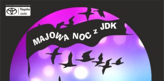 Śpiew, taniec, muzyka, teatr: Majowa Noc z JDK baner na czarnym tle fioletowo granatowe koło. Na nim biały napis na czarnej fladze, majowa noc z jdk, pod nią czarne ptaki, na końcu flagi przyczepiona para jedzie na rowerze, ciągnie za sobą wózek z klatką. . Pod kołem białym kolorem napisana data.