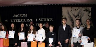 Na tle baneru konkursowego sacrum w literaturze stoją nagrodzeni i wyróżnieni młodzi uczestnicy. W rękach trzymają papierowe torby oraz dyplomy