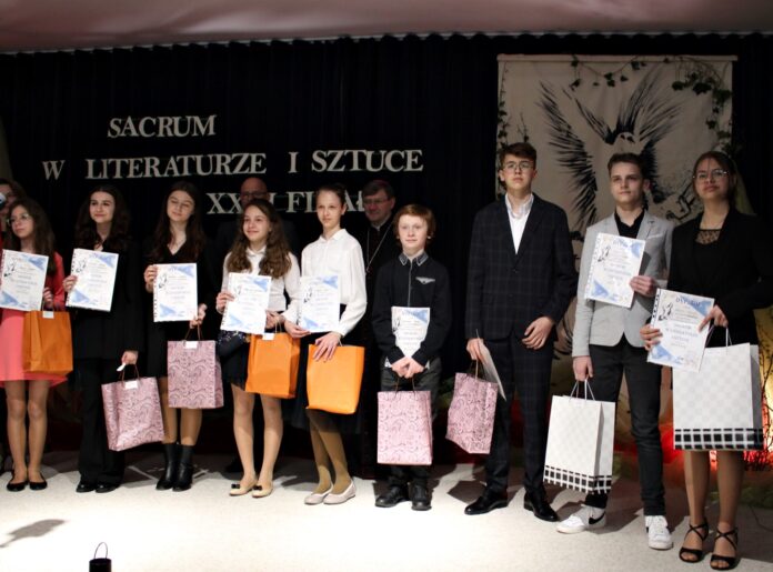 tymek Na tle baneru konkursowego sacrum w literaturze stoją nagrodzeni i wyróżnieni młodzi uczestnicy. W rękach trzymają papierowe torby oraz dyplomy
