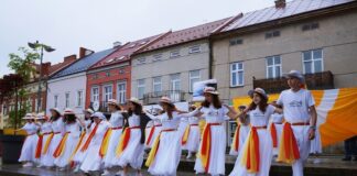 Grupa młodzieży wraz z siostrą zakonną – Kariną Pawłowską tańczy w strugach deszczu. Wszyscy ubrani są na biało. Każda z osób ma przywiązaną chustę w kolorze żółto-czerwonym.