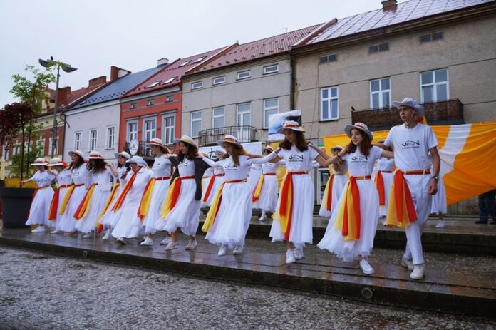 1 Grupa młodzieży wraz z siostrą zakonną – Kariną Pawłowską tańczy w strugach deszczu. Wszyscy ubrani są na biało. Każda z osób ma przywiązaną chustę w kolorze żółto-czerwonym.