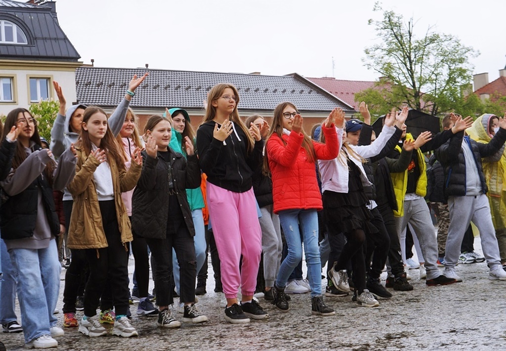 Grupa radosnych dzieci, tańczących w strugach deszczu na płycie jasielskiego Rynku. Wszyscy unoszą ręce w górę. 