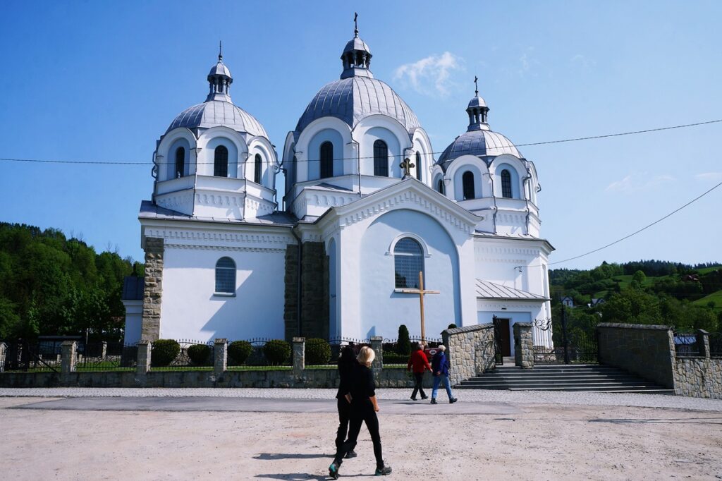 Uczestnicy wycieczki zmierzają w stronę Cerkwi Opieki Matki Bożej w Szlachtowej. Świątynia jest duża. Na zewnątrz w kolorze biało – błękitnym.