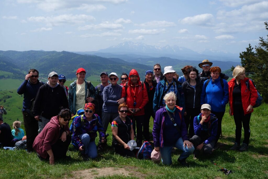 Uczestnicy wycieczki na szczycie Wysoki Wierch w otoczeniu górskich wzniesień, lasów i Tatr pokrytych białym śniegiem. 