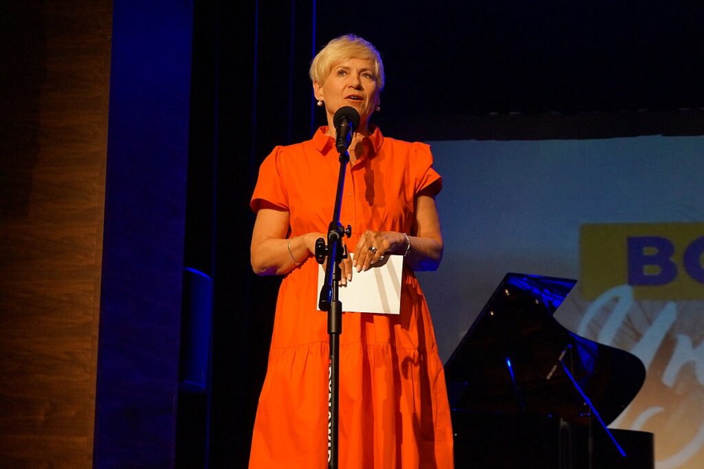 Na scenie Pani Dyrektor Jasielskiego Domu Kultury – Helena Gołębiowska, wita zgromadzoną publiczność. Ma na sobie długą, pomarańczową sukienkę. W oddali stoi czarny fortepian.