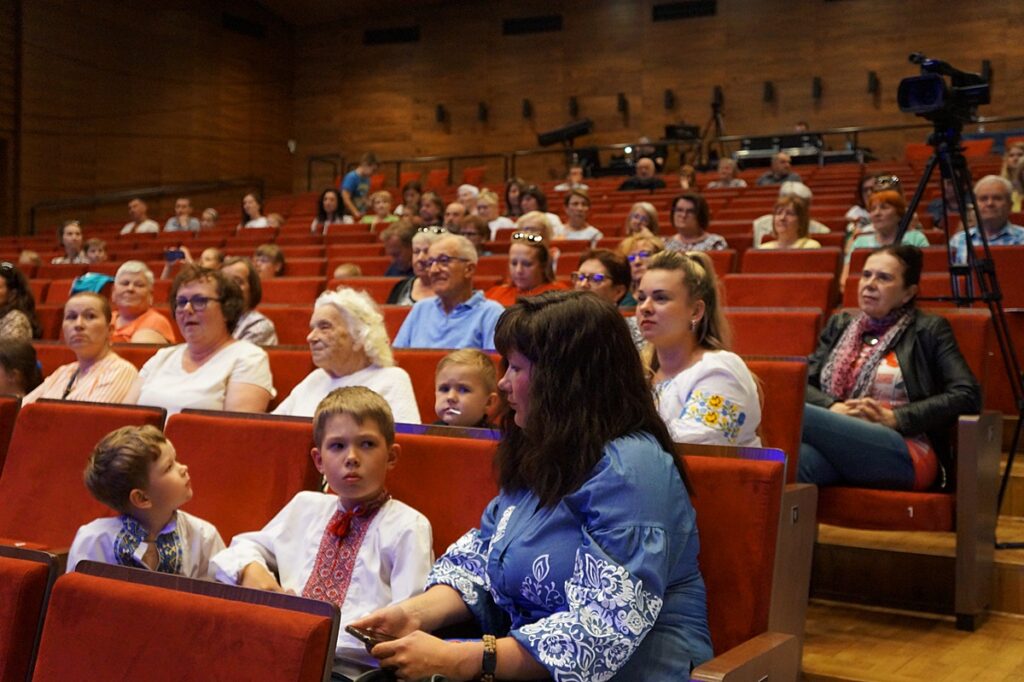Publiczność w radosnym oczekiwaniu na występ. Na sali widowiskowej są obywatele Ukrainy. Ubrani są w kolorowe koszule z naszyciami.