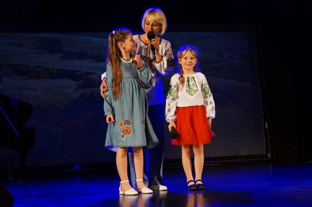 Trzy osoby na scenie. Kobieta i dwie dziewczynki. Każda ubrana jest w strój w ukraińskie wzory. Kobieta zapowiada występ dziewczynek.