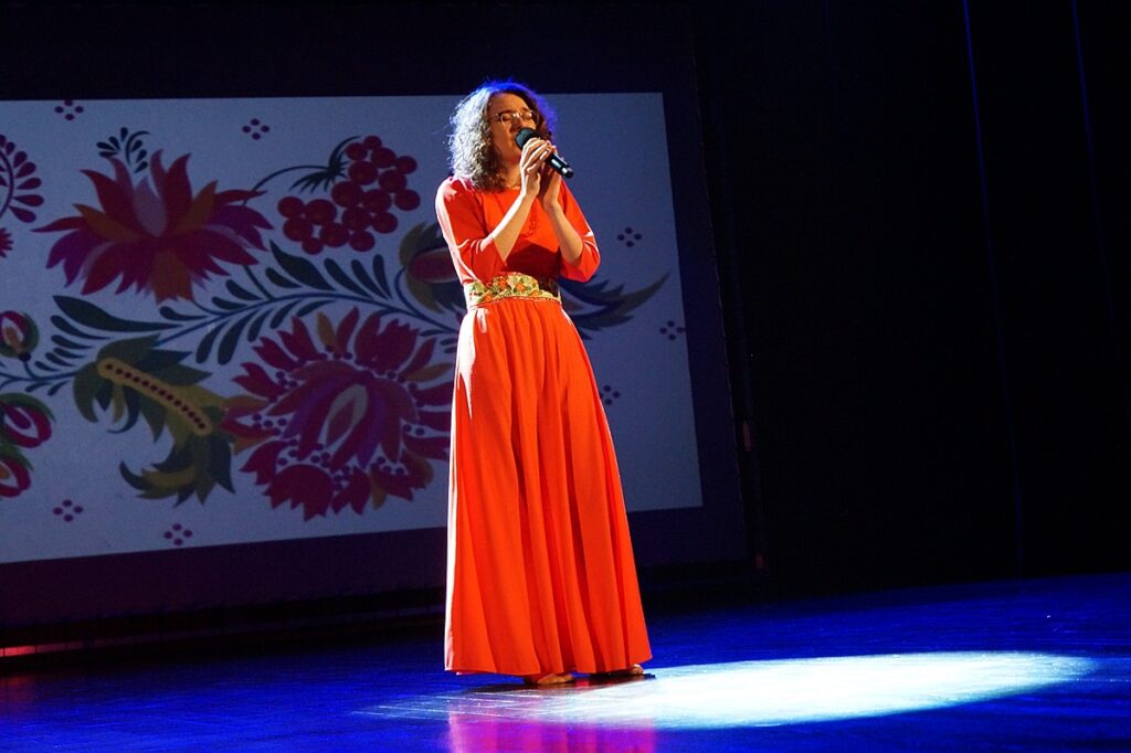 Młoda dziewczyna śpiewa na scenie. Ubrana jest w długą, pomarańczową suknię z ozdobnym pasem w ukraińskie wzorki.