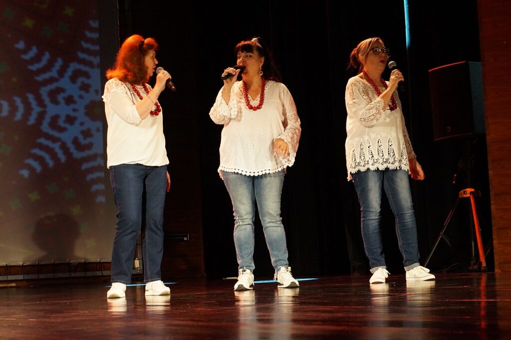 Trzy kobiety na scenie śpiewają piosenkę. Każda ubrana jest w białą bluzkę, jasne spodnie i buty. Elementem dodatkowym stroju są czerwone korale.