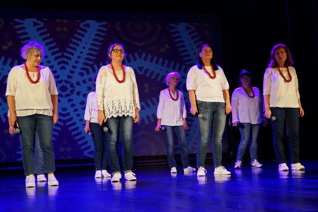 Grupa kobiet na scenie. Ubrane są w białe bluzki, jasne spodnie i buty. Elementem dodatkowym stroju są czerwone korale.