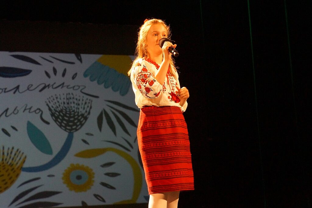 Dziewczynka na scenie śpiewa piosenkę. Ubrana jest w piękny strój ukraiński z naszyciami.