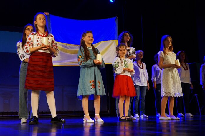 DSC03756 Na scenie dwie młode dziewczyny z flagą Ukrainy przed, którą stoją cztery dziewczynki, trzymające w dłoniach świece.