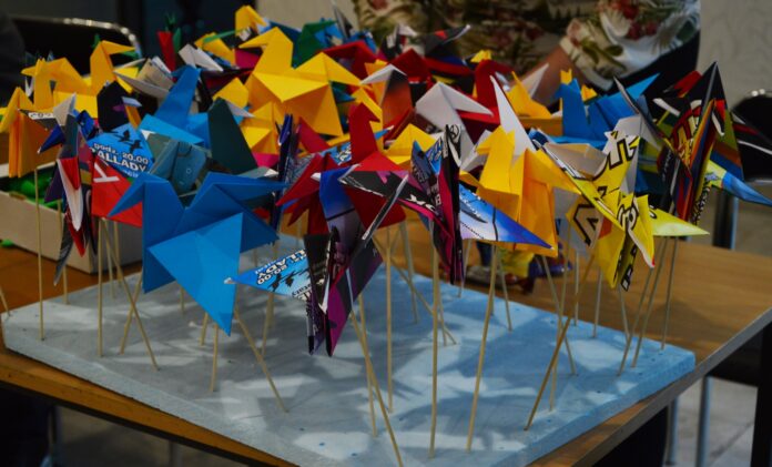 DSC_3264 Na stole leży duży kawałek styropianu w kształcie prostokąta. do niego są wpięte na patykach kolorowe żurawie