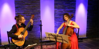 Spod znaku gitary i wiolonczeli Obraz zawiera: Dwoje młodych ludzi - mężczyzna i kobieta siedzą twarzami zwróconymi do siebie. Mężczyzna gra na gitarze, kobieta na wiolonczeli. Przed każdym z nich stoi pulpit nutowy. Pomiędzy nimi statyw z mikrofonem. Pod nogami maja wyłożony dywan.