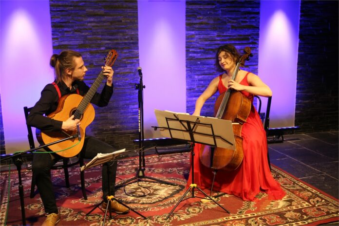 Obraz zawiera: Dwoje młodych ludzi - mężczyzna i kobieta siedzą twarzami zwróconymi do siebie. Mężczyzna gra na gitarze, kobieta na wiolonczeli. Przed każdym z nich stoi pulpit nutowy. Pomiędzy nimi statyw z mikrofonem. Pod nogami maja wyłożony dywan.