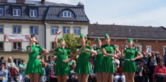 Dzień Godności Osób z Niepełnosprawnością Intelektualną Obraz zawiera: Osiem mażoretek w dwóch rzędach tańczy z batutami na płycie jasielskiego rynku. Z tyłu podopieczni warsztatów terapii zajęciowej ze Specjalnego Ośrodka Szkolno - Wychowawczego w Jaśle i innych ośrodków. W tle budynki Starostwa Powiatowego w Jaśle oraz z prawej strony niższy, jednopiętrowy budynek w którym mieści się biuro europosła Bogdana Rzońcy.