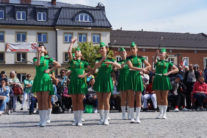 Obraz zawiera: Osiem mażoretek w dwóch rzędach tańczy z batutami na płycie jasielskiego rynku. Z tyłu podopieczni warsztatów terapii zajęciowej ze Specjalnego Ośrodka Szkolno - Wychowawczego w Jaśle i innych ośrodków. W tle budynki Starostwa Powiatowego w Jaśle oraz z prawej strony niższy, jednopiętrowy budynek w którym mieści się biuro europosła Bogdana Rzońcy.