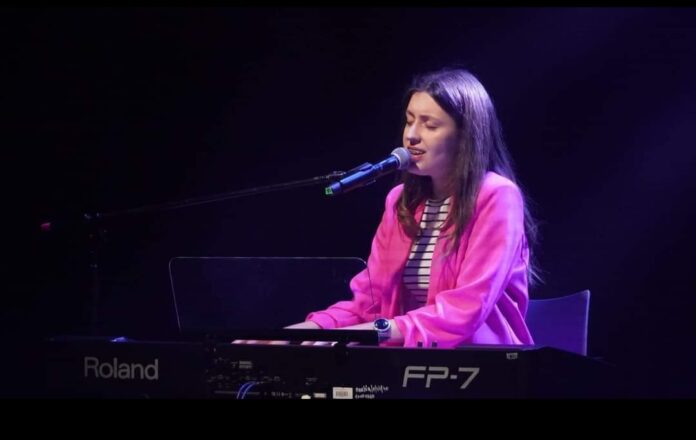 Karolina Polak w Opolu Przy kejbordzie siedzi Karolina Polak podczas swojego występu. Nastolatka ma ciemne, długie włosy, ubrana jest w różową marynarkę oraz białą bluzkę w czarne cienkie paski. Przed nią mikrofon, do którego śpiewa.