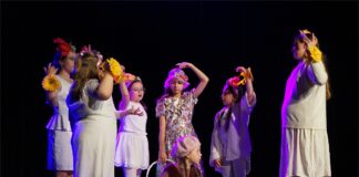 Obraz zawiera: Na zdjęcu widać grupę dzieci: 7 dziewczynek. Jedna z nich to główna bohaterka – młodziutka Gerda, która zmierza świat w poszukiwaniu Kaja. Dziewczynka przebywa w otoczeniu radosnych przyjaciółek. Pięć dziewczynek ubranych jest w stroje koloru białego. Dodatkowym elementem ubioru każdej z nich są żółte kwiaty na dłoniach, symbolizujące nadzieję i radość. Jedna z dziewczynek ubrana jest w kremową sukienkę z motywem czerwonych kwiatów i biały kapelusz z czerwoną różą. W prawej dłoni trzyma biały koszyk. Z kolei Gerda znajduje się w pozycji siedzącej. Ubrana jest w jasnobrązowy sweterek, ciemnobrązowy kożuszek i brązową spódniczkę. Ma długie, jasne warkocze, a na jej głowie beżowy berecik z dodatkiem pereł. Scena rozgrywa się w ciemno-jasnym oświetleniu.