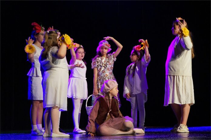 Królowa Śniegu3_3 Obraz zawiera: Na zdjęcu widać grupę dzieci: 7 dziewczynek. Jedna z nich to główna bohaterka – młodziutka Gerda, która zmierza świat w poszukiwaniu Kaja. Dziewczynka przebywa w otoczeniu radosnych przyjaciółek. Pięć dziewczynek ubranych jest w stroje koloru białego. Dodatkowym elementem ubioru każdej z nich są żółte kwiaty na dłoniach, symbolizujące nadzieję i radość. Jedna z dziewczynek ubrana jest w kremową sukienkę z motywem czerwonych kwiatów i biały kapelusz z czerwoną różą. W prawej dłoni trzyma biały koszyk. Z kolei Gerda znajduje się w pozycji siedzącej. Ubrana jest w jasnobrązowy sweterek, ciemnobrązowy kożuszek i brązową spódniczkę. Ma długie, jasne warkocze, a na jej głowie beżowy berecik z dodatkiem pereł. Scena rozgrywa się w ciemno-jasnym oświetleniu.