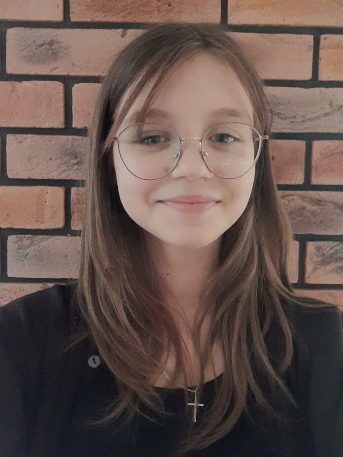 Matylda Obraz zawiera: Kilkunastoletnia uśmiechnięta dziewczynka w okularach z ciemnymi, długimi włosami sięgającymi nieco poza ramiona. Ubrana w ciemny żakiet. Na szyi ma łańcuszek z krzyżykiem. Stoi na tle ceglanego muru.