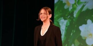 „Pamiętajcie o ogrodach” – nagrody dla Emilii Starzec i Natalii Żegleń Obraz zawiera: Młoda kobieta w okularach ze związanymi z tyłu włosami stoi na scenie. Ubrana jest w czarną sukienkę mini i w czarny żakiet. Na szyi ma łańcuszek. Ręce trzyma za sobą. Z tyłu, na dalszym planie widać ekran na którym wyświetlane jest zdjęcie kwiatów jabłoni.