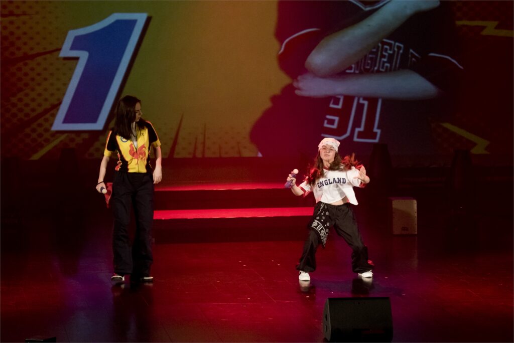 Obraz zawiera: Dwie młode dziewczyny na scenie JDK Pierwsza z lwej trzyma w dłoniach atrapę mikrofonu,w sportowej, rozsuwanej bluzie z krótkim rękawem i w sportowych spodniach spogląda w kierunku znajdującej się po prawej stronie drugiej dziewczyny, która wykonuje taneczne figury hip - hopowe. W prawej ręce trzyma atrapę mikrofonu i przechyla się do tyłu.