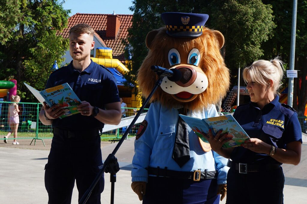 Trzy osoby: mężczyzna (policjant), kobieta (Straż Miejska) i policyjny miś, czytają ,Lokomotywę’’ Juliana Tuwima.