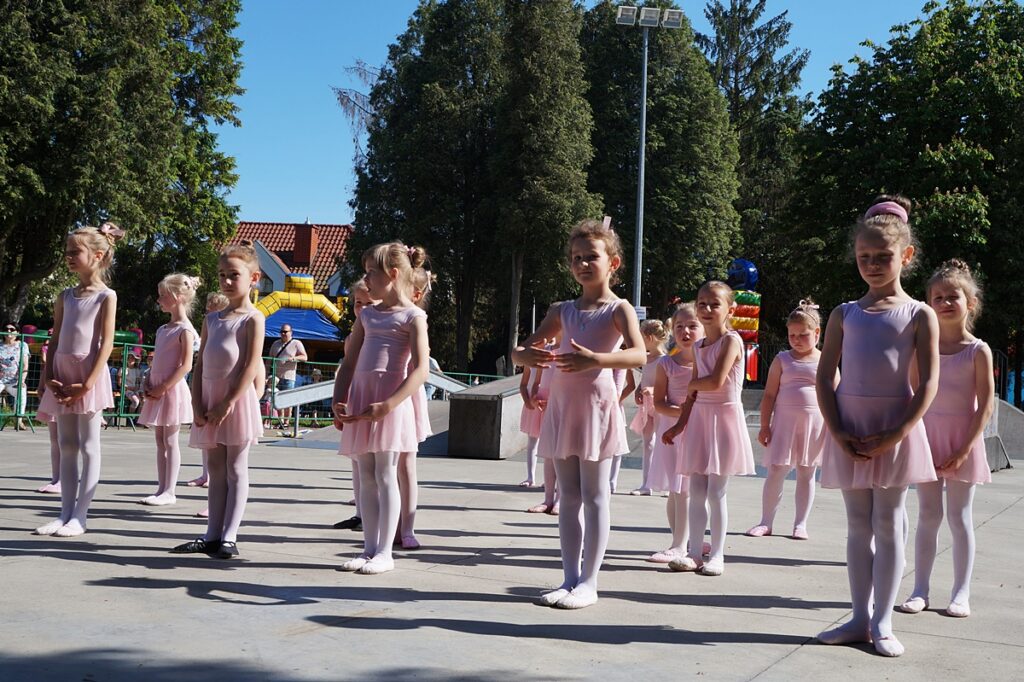 Grupa małych dziewczynek tańczy układ baletowy. Wszystkie ubrane są w strój koloru jasno różowego. 