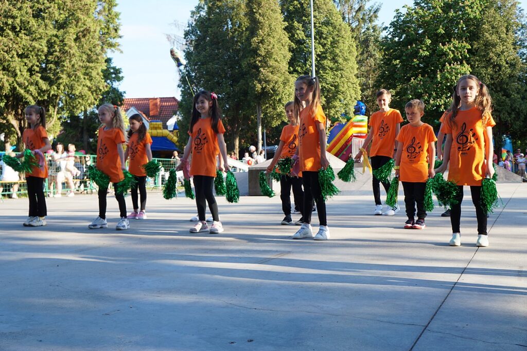 Grupa dzieci wykonuje układ taneczny. Każde dziecko ubrane jest w pomarańczową koszulkę. 