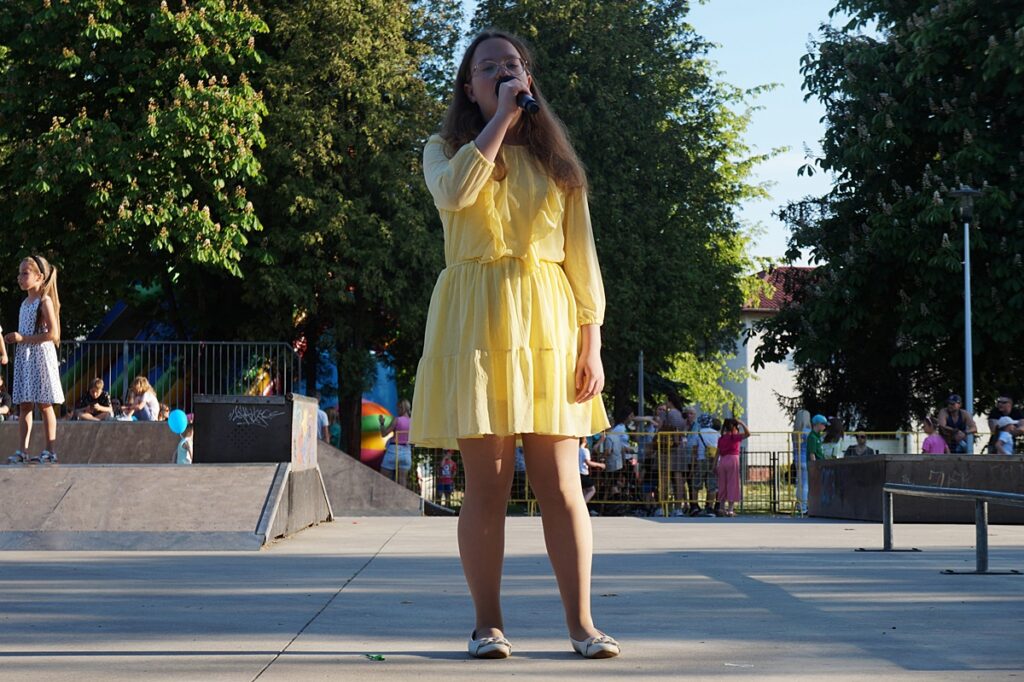 Młoda dziewczyna śpiewa piosenkę. Ubrana jest w żółtą sukienkę. 