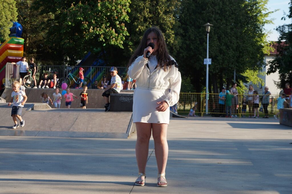 Młoda dziewczyna śpiewa piosenkę. Ubrana jest w białą spódniczkę i kurtkę. 
