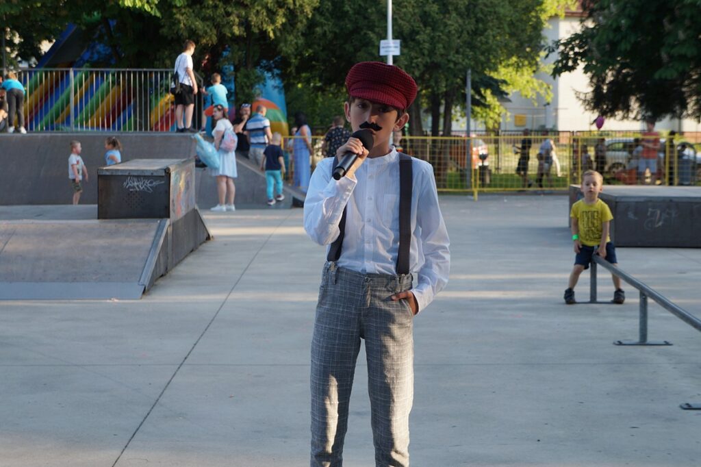 Młody chłopiec śpiewa piosenkę. Ubrany jest w czerwony beret z daszkiem, białą koszulę i szare spodnie w białą kratkę.