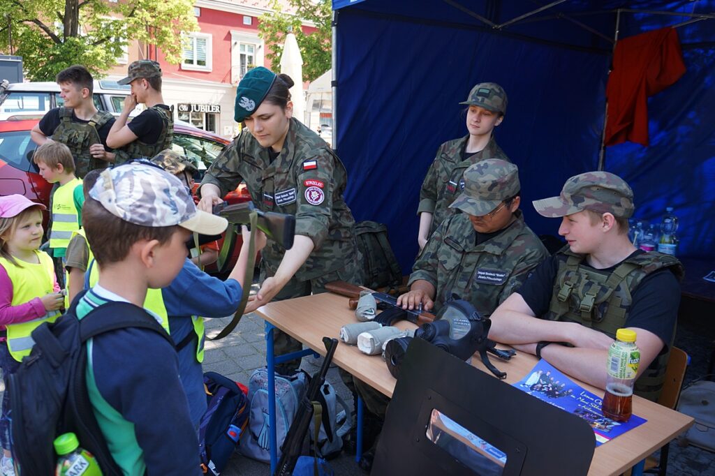 Młodzi żołnierze przy specjalnym stoisku prezentują dzieciom sprzęt wojskowy: karabin i maskę tlenową.