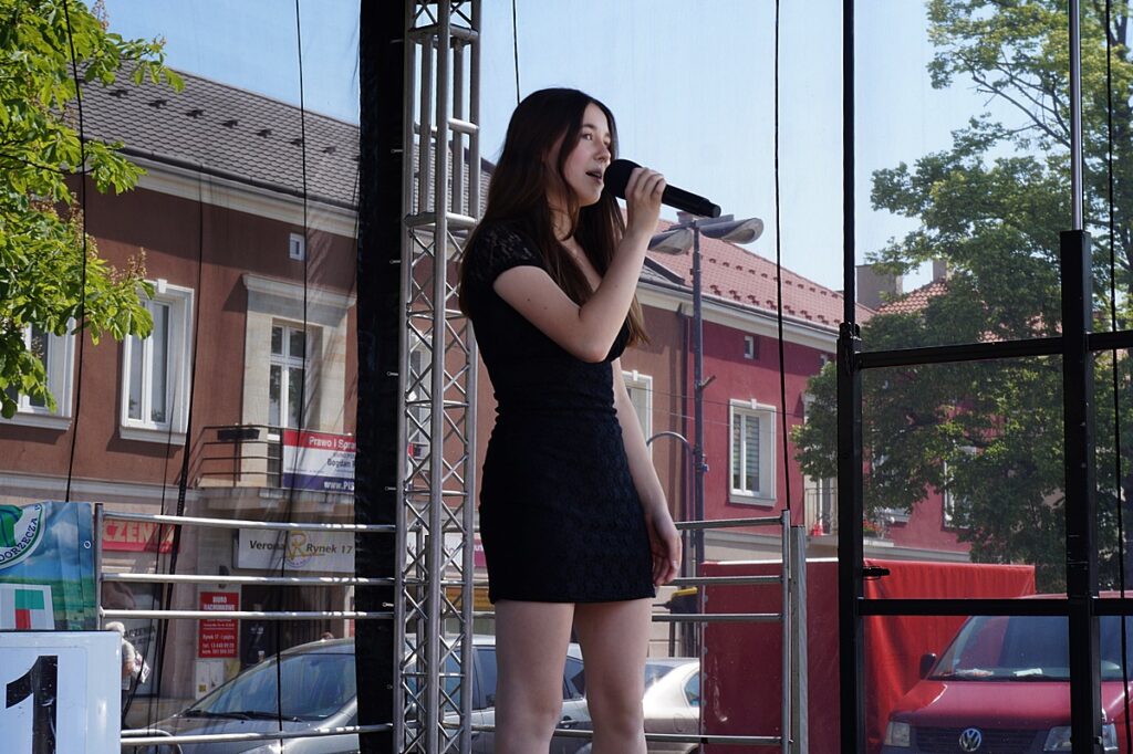 Młoda dziewczyna na scenie śpiewa piosenkę. Ubrana jest w czarną sukienkę. W ręku trzyma mikrofon. 
