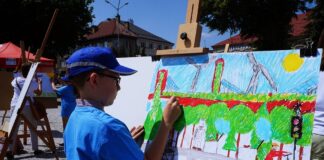 Artystycznie, słonecznie i kolorowo Chłopiec maluje obraz. Ubrany jest w niebieską czapkę z daszkiem z szarym paskiem i niebieską koszulkę. Maluje zielony las, drogę, ludzi, niebieskie niebo, słońce i wiatraki ekologiczne na trawie.