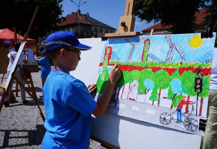 Chłopiec maluje obraz. Ubrany jest w niebieską czapkę z daszkiem z szarym paskiem i niebieską koszulkę. Maluje zielony las, drogę, ludzi, niebieskie niebo, słońce i wiatraki ekologiczne na trawie.