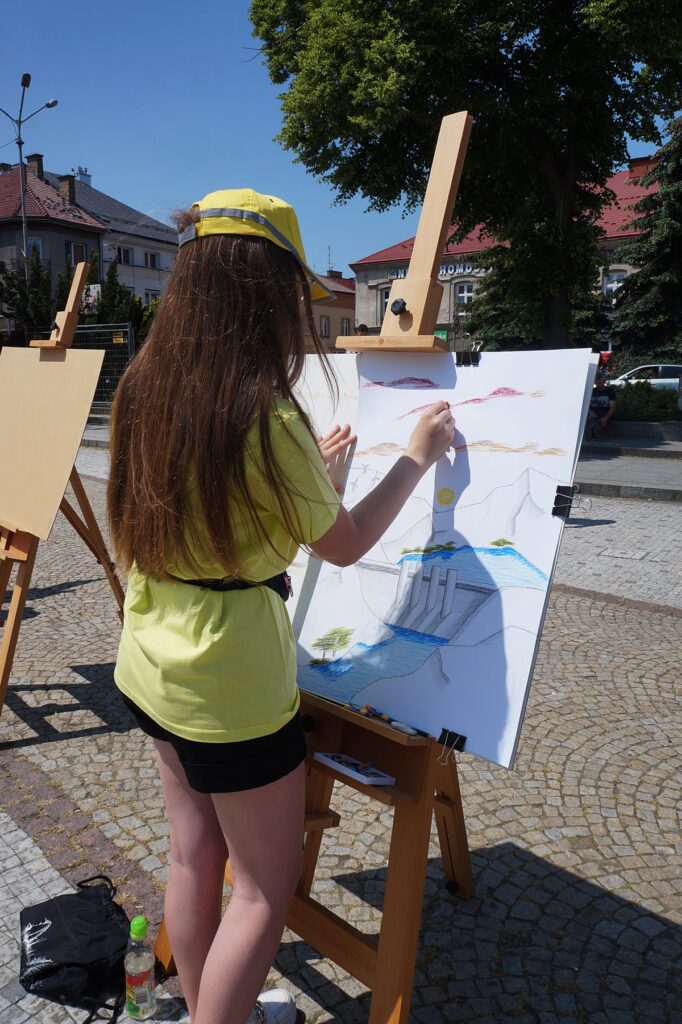 Dziewczynka maluje obraz. Ubrana jest w żółtą koszulkę, żółtą czapkę z daszkiem z szarym paskiem i czarne krótkie spodenki. Dziecko szkicuje góry. 