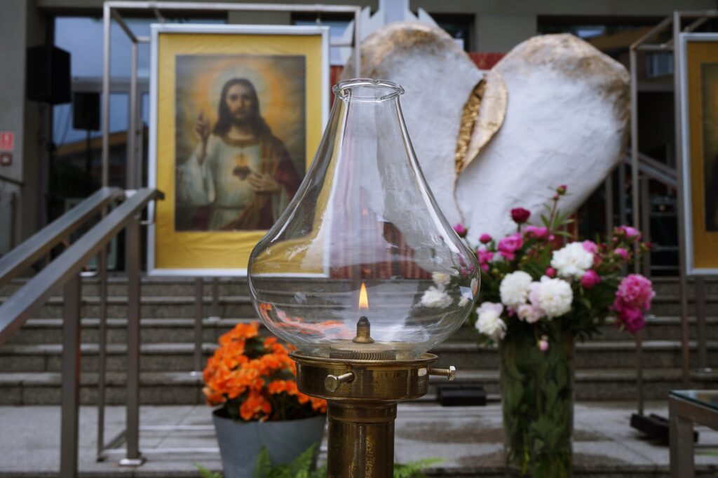 Świecznik liturgiczny z płomieniem. W oddali stoi duże, białe serce i obraz Najświętszego Serca Pana Jezusa. 
